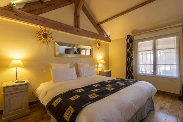 Chambre Amazone : lit sous la charpente, jeté sombre à motifs dorés, miroirs soleil et fenêtre