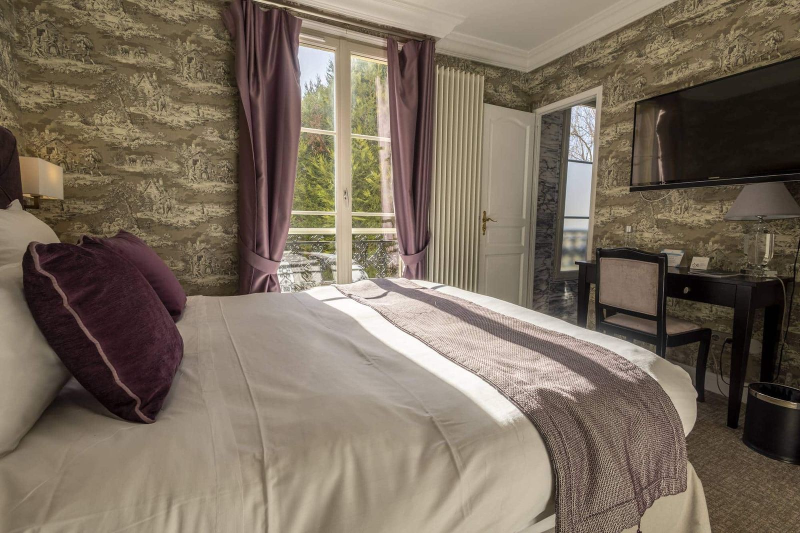 Chambre Coquillage : lit aux coussins prune, rideaux mauves et fenêtre sur les arbres