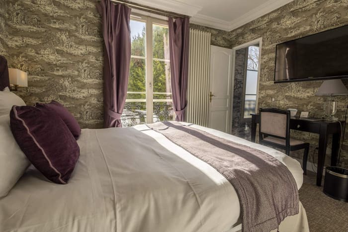 Chambre Coquillage : lit aux coussins prune, rideaux mauves et fenêtre sur les arbres