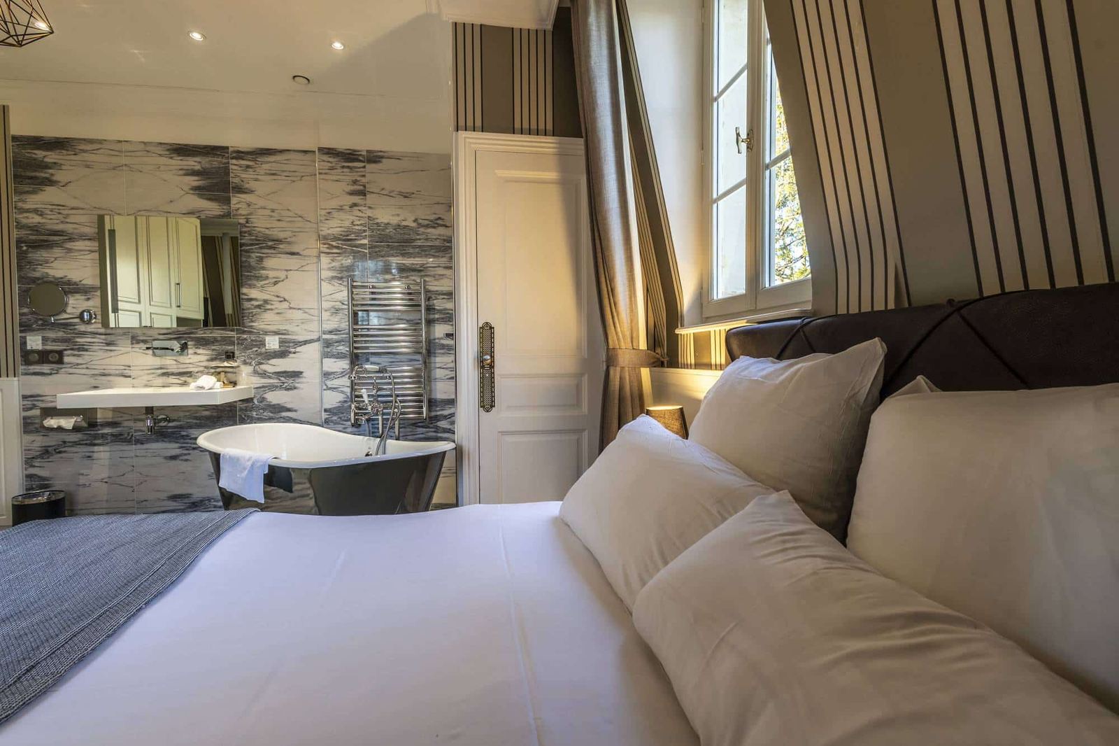 Chambre Coquine : lit au premier plan, baignoire sur pieds et lavabo sur mur de marbre, fenêtre
