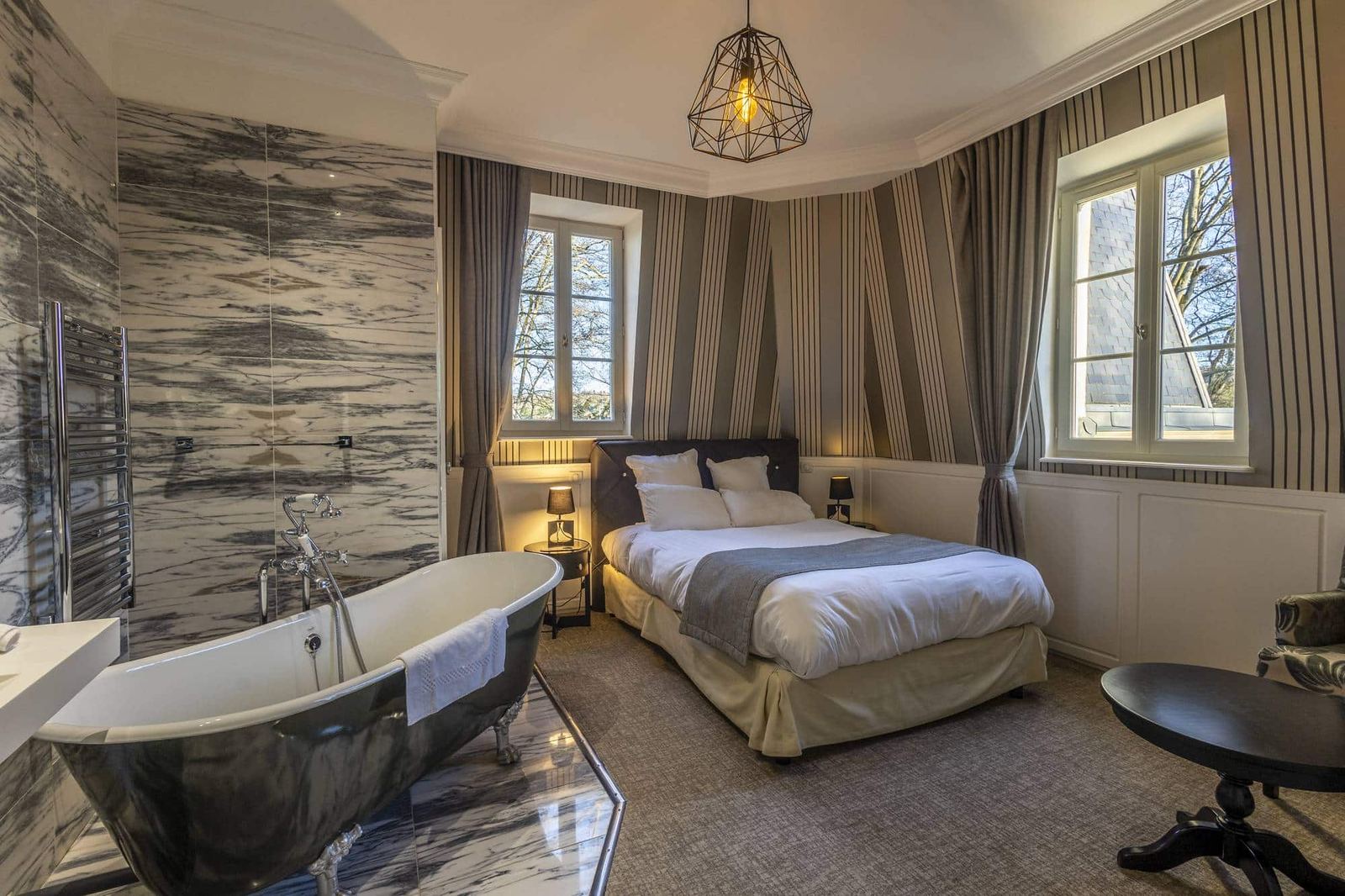 Chambre Coquine : baignoire sur pieds, mur de marbre, lit et deux fenêtres sur les arbres