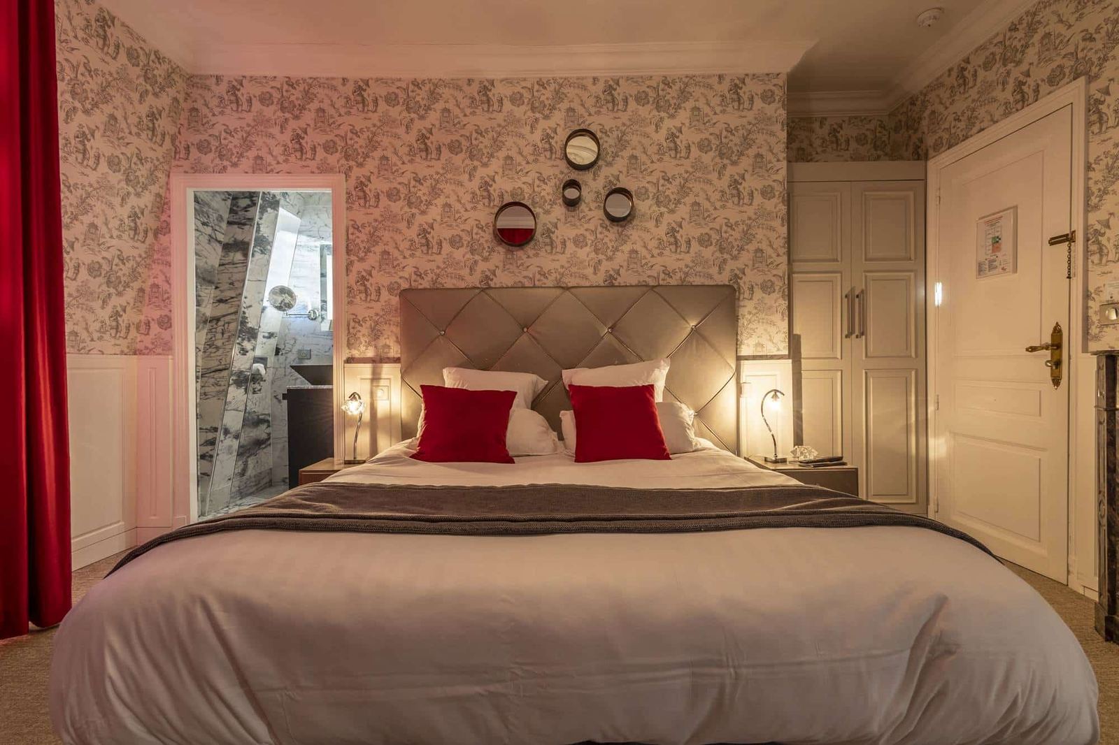 Chambre Dorée : lit capitonné vu de face, coussins rouges, toile de Jouy et miroirs ronds
