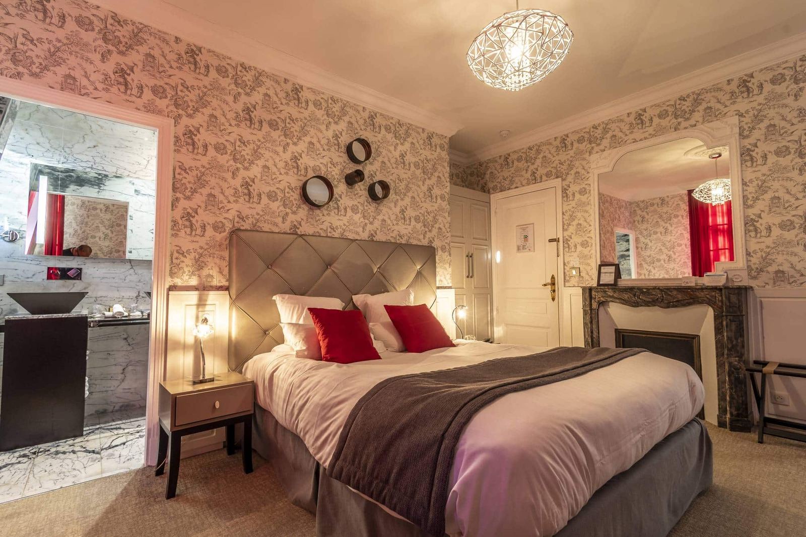 Chambre Dorée : lit capitonné, coussins rouges, cheminée de marbre et suspension ajourée