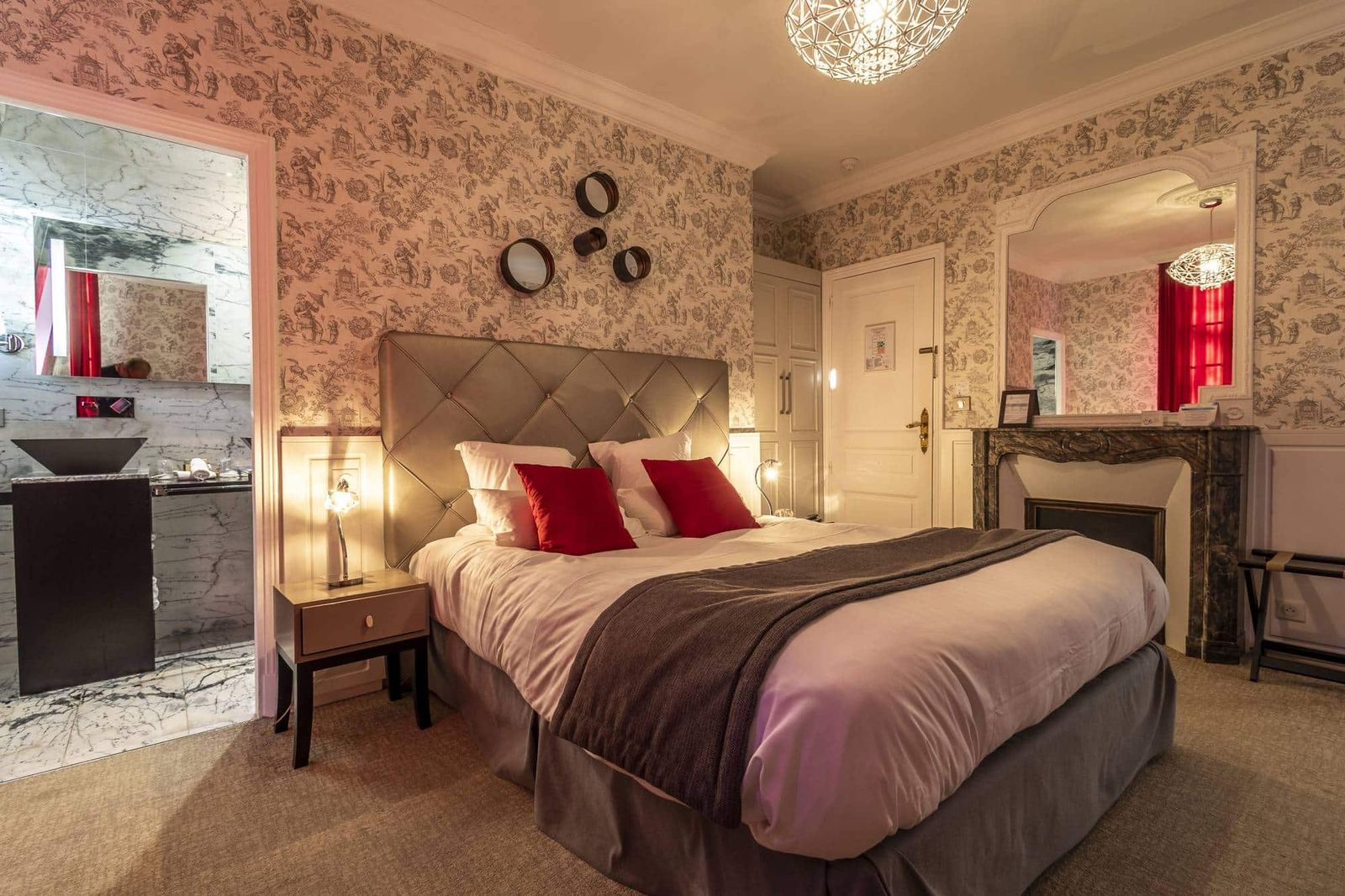 Chambre Dorée : lit capitonné, coussins rouges et cheminée de marbre sous une lumière chaude