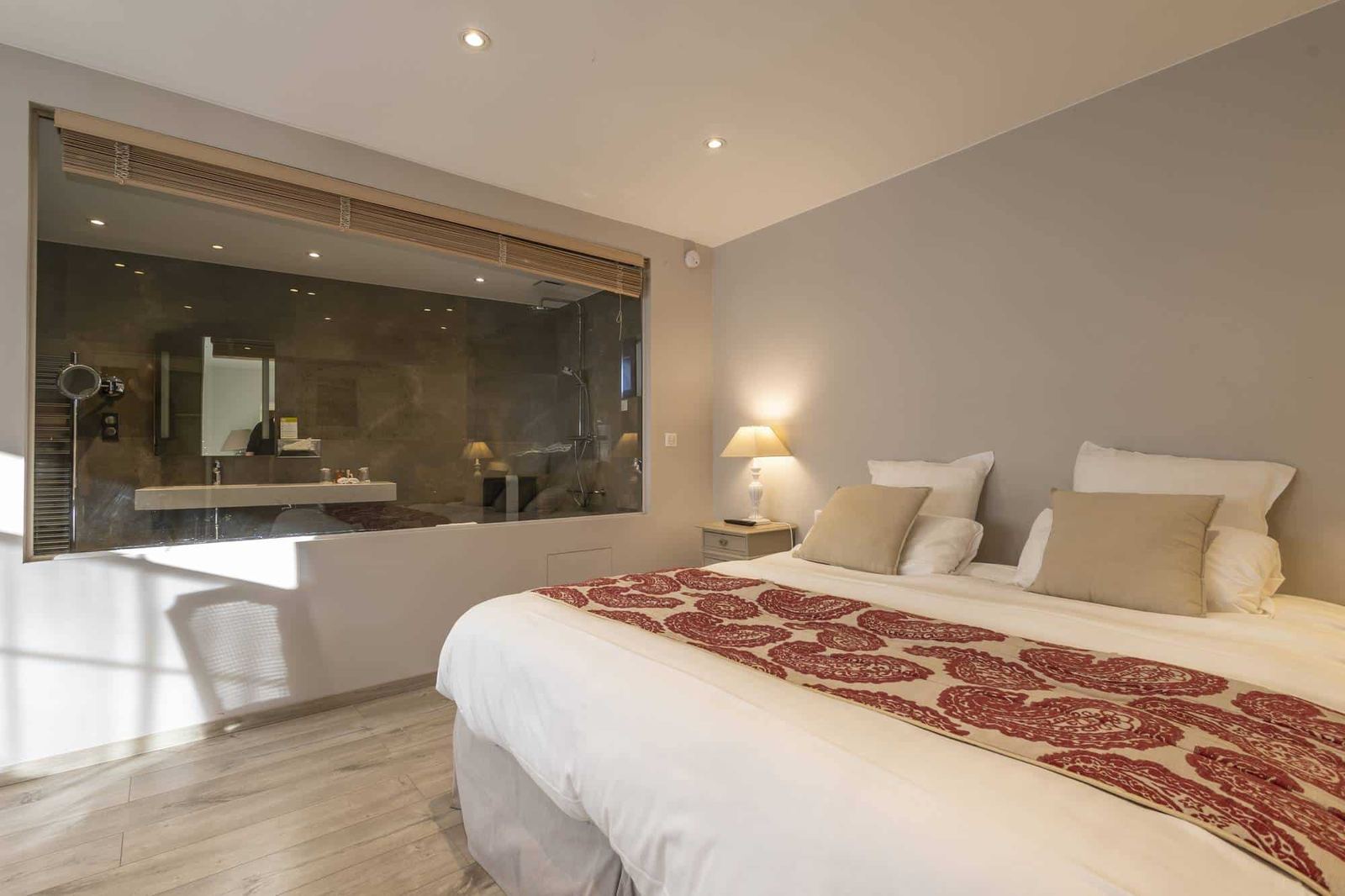 Chambre Centaure : lit au chemin de lit rouge, vitre ouverte sur la douche et parquet clair