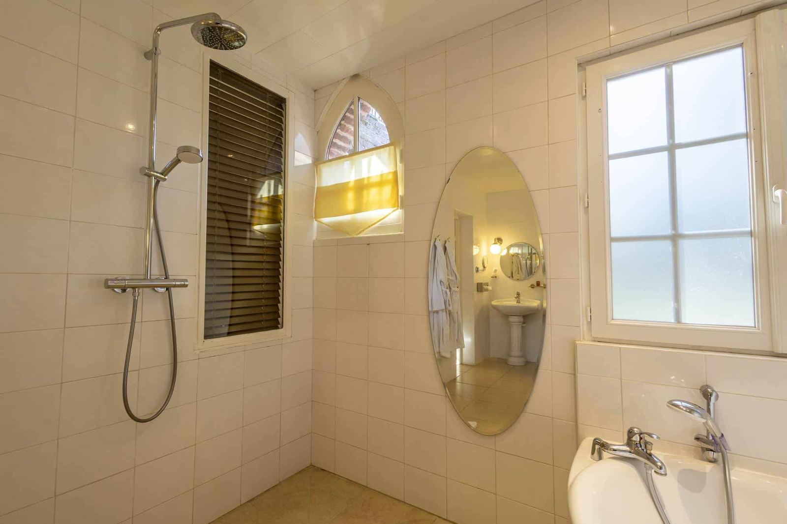Chambre Poulinière : colonne de douche, miroir ovale, carrelage blanc et petite fenêtre en ogive
