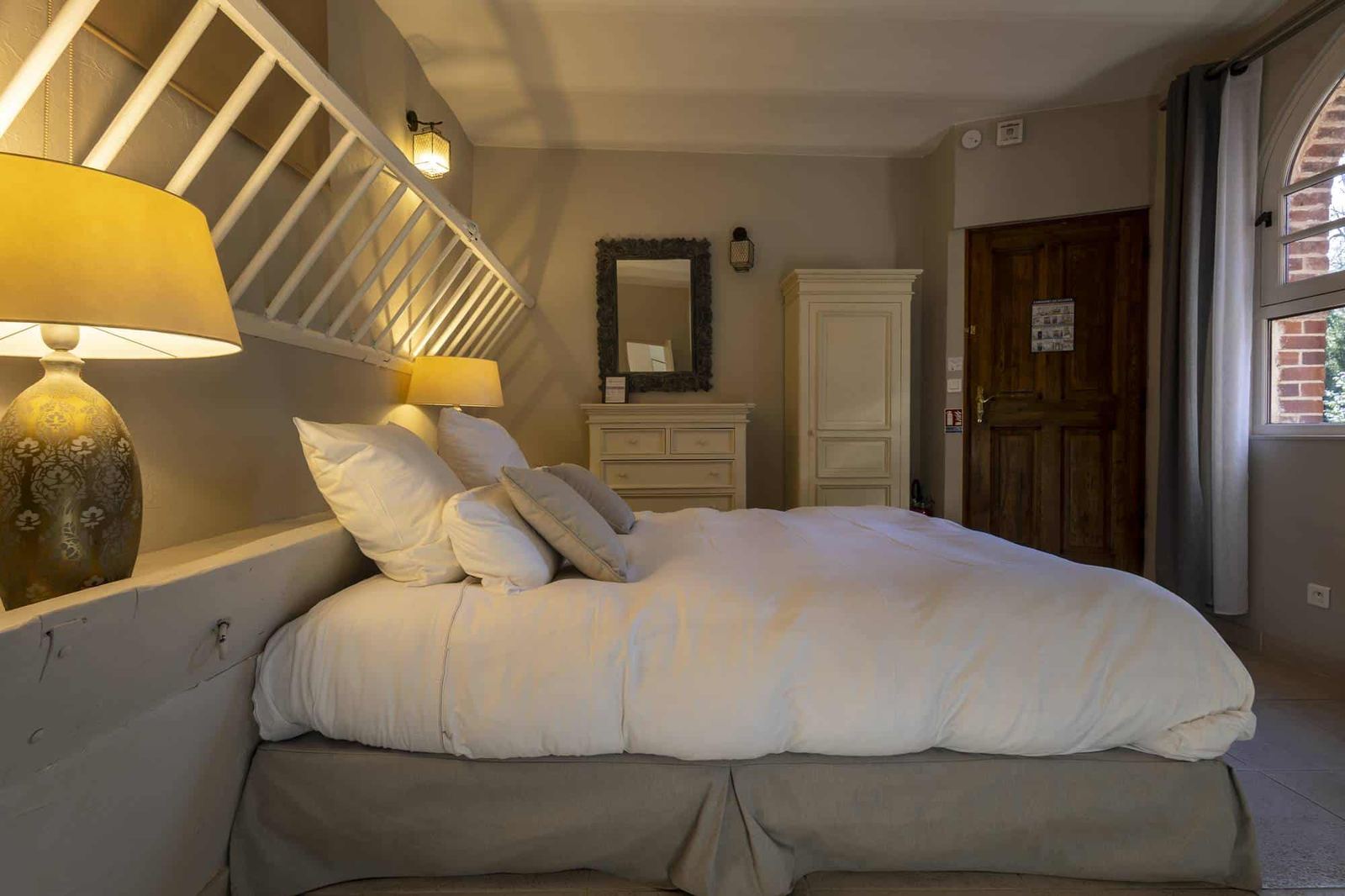Chambre Poulinière : lit aux oreillers blancs, grande lampe dorée, commode, armoire et porte en bois