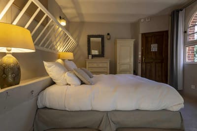 Chambre Poulinière : lit aux oreillers blancs, grande lampe dorée, commode, armoire et porte en bois