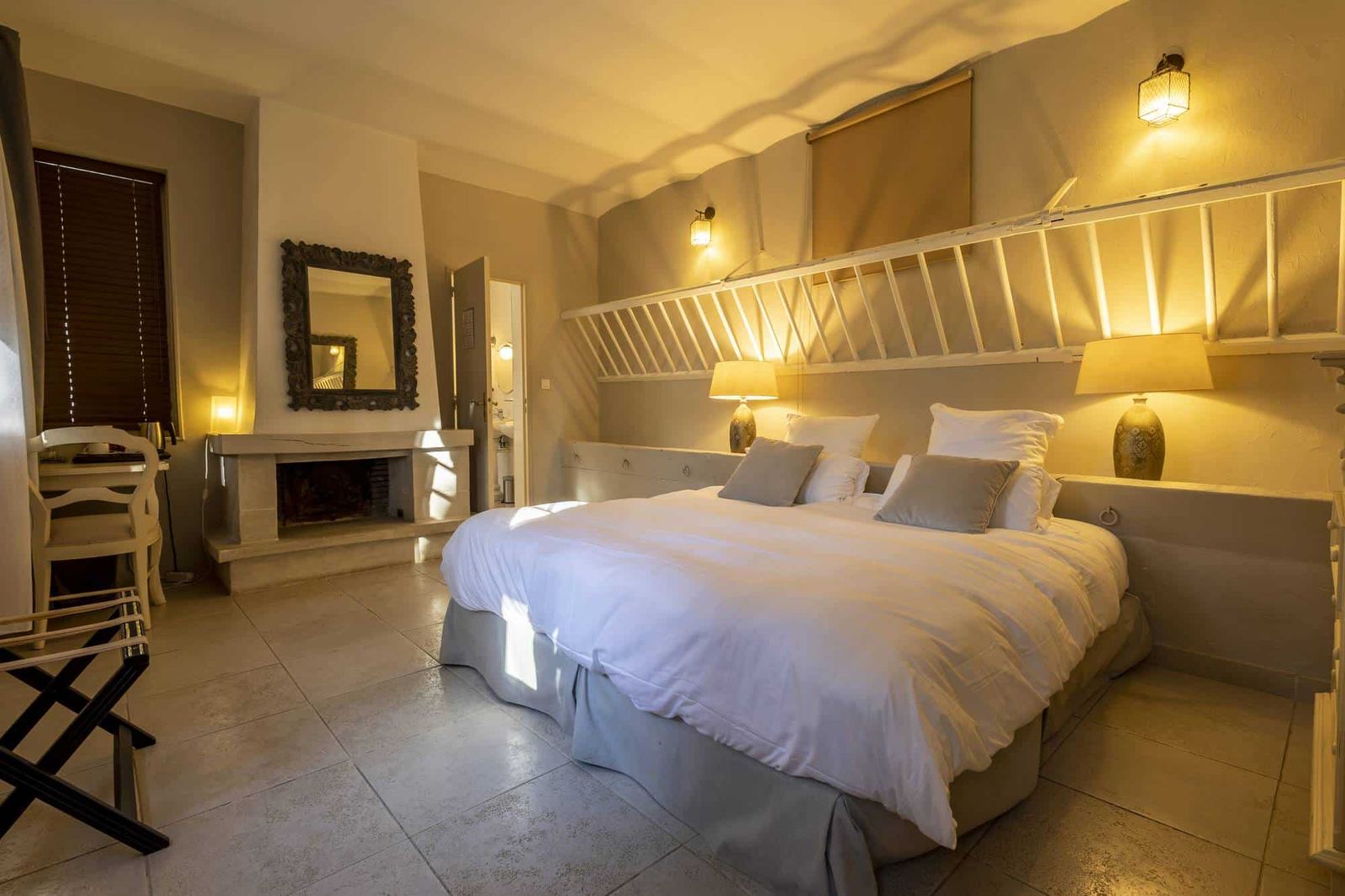 Chambre Poulinière : lit blanc sous un râtelier en bois blanc, cheminée en pierre et lampes dorées