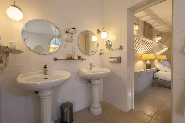 Chambre Poulinière : deux lavabos sur colonne, miroirs ronds et carrelage blanc, ouverture sur le lit