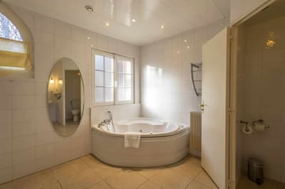 Chambre Poulinière : baignoire d’angle à jets, carrelage blanc, miroir ovale et fenêtre