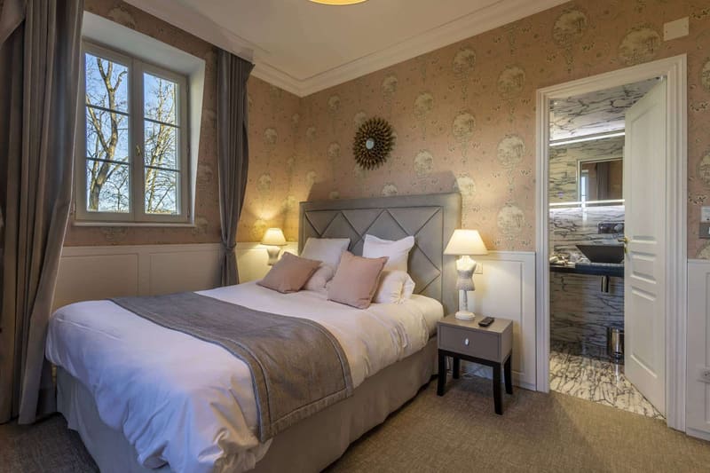 Chambre Romantique : lit aux coussins rose poudré, papier peint à médaillons et fenêtre sur les arbres