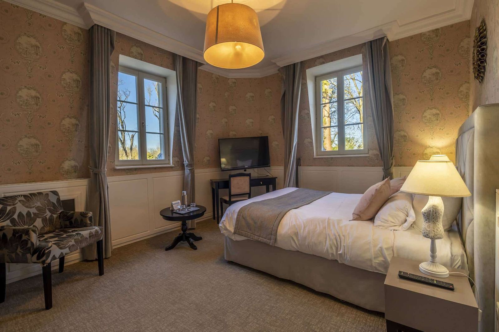 Chambre Romantique : lit, fauteuil fleuri et deux fenêtres sur les arbres, murs rose poudré