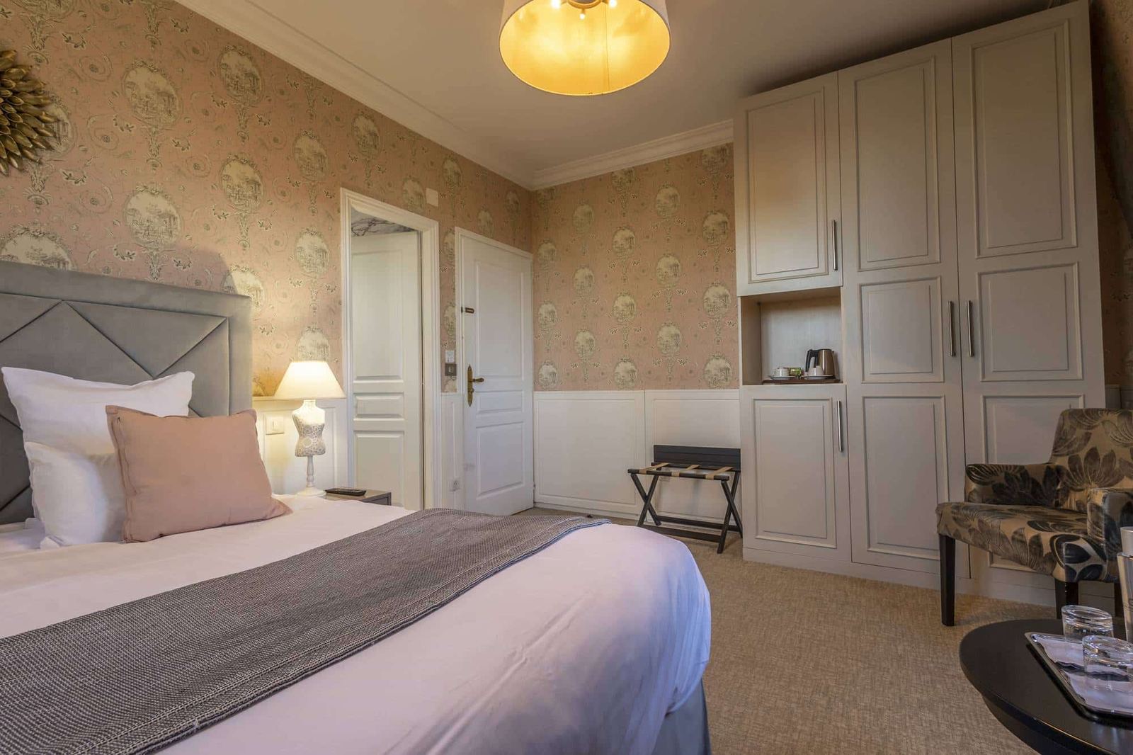 Chambre Romantique : lit, grande armoire gris clair et fauteuil fleuri sous un plafonnier doré