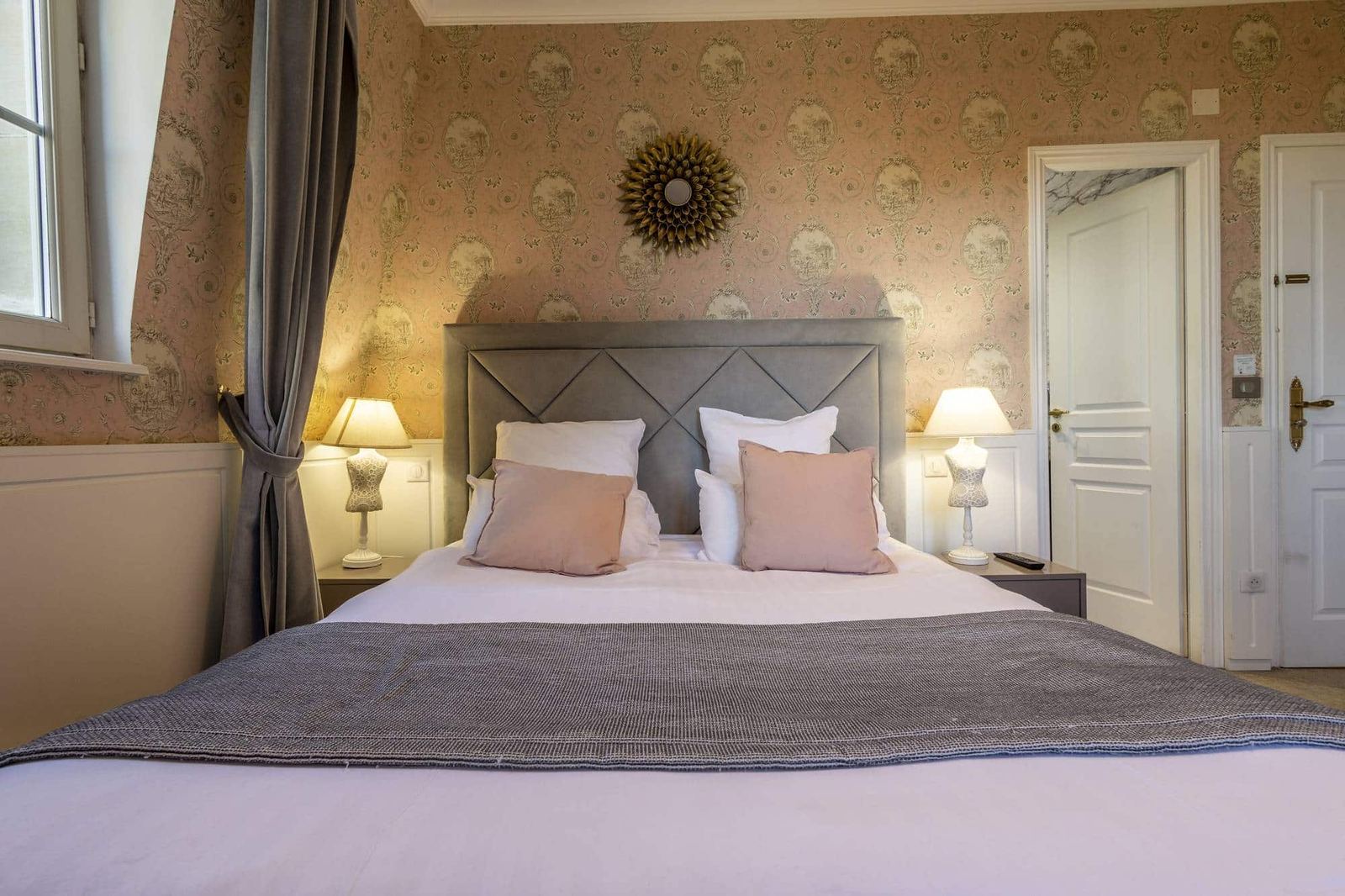Chambre Romantique : lit sous un miroir soleil doré, coussins rose poudré et deux lampes de chevet