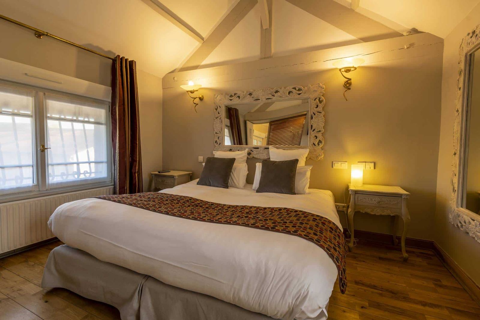Chambre Voltige : lit vu de face sous les poutres, miroir baroque blanc et appliques allumées