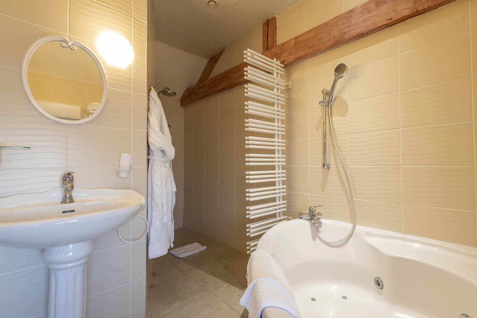 Chambre Voltige : salle de bain crème sous poutres, baignoire d’angle à jets et lavabo colonne