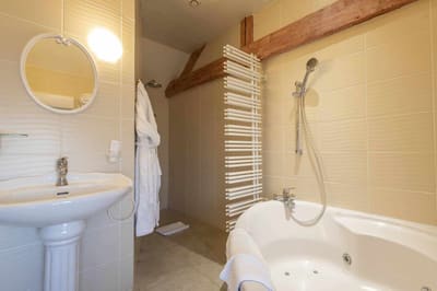 Chambre Voltige : salle de bain crème sous poutres, baignoire d’angle à jets et lavabo colonne