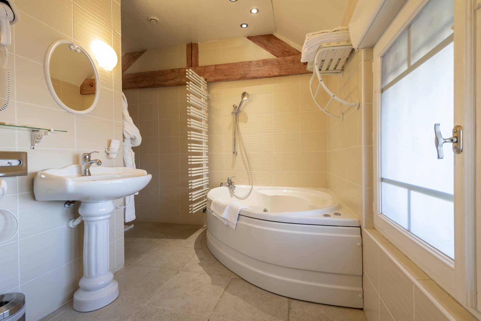 Chambre Voltige : baignoire d’angle près d’une fenêtre dépolie, lavabo colonne et poutres