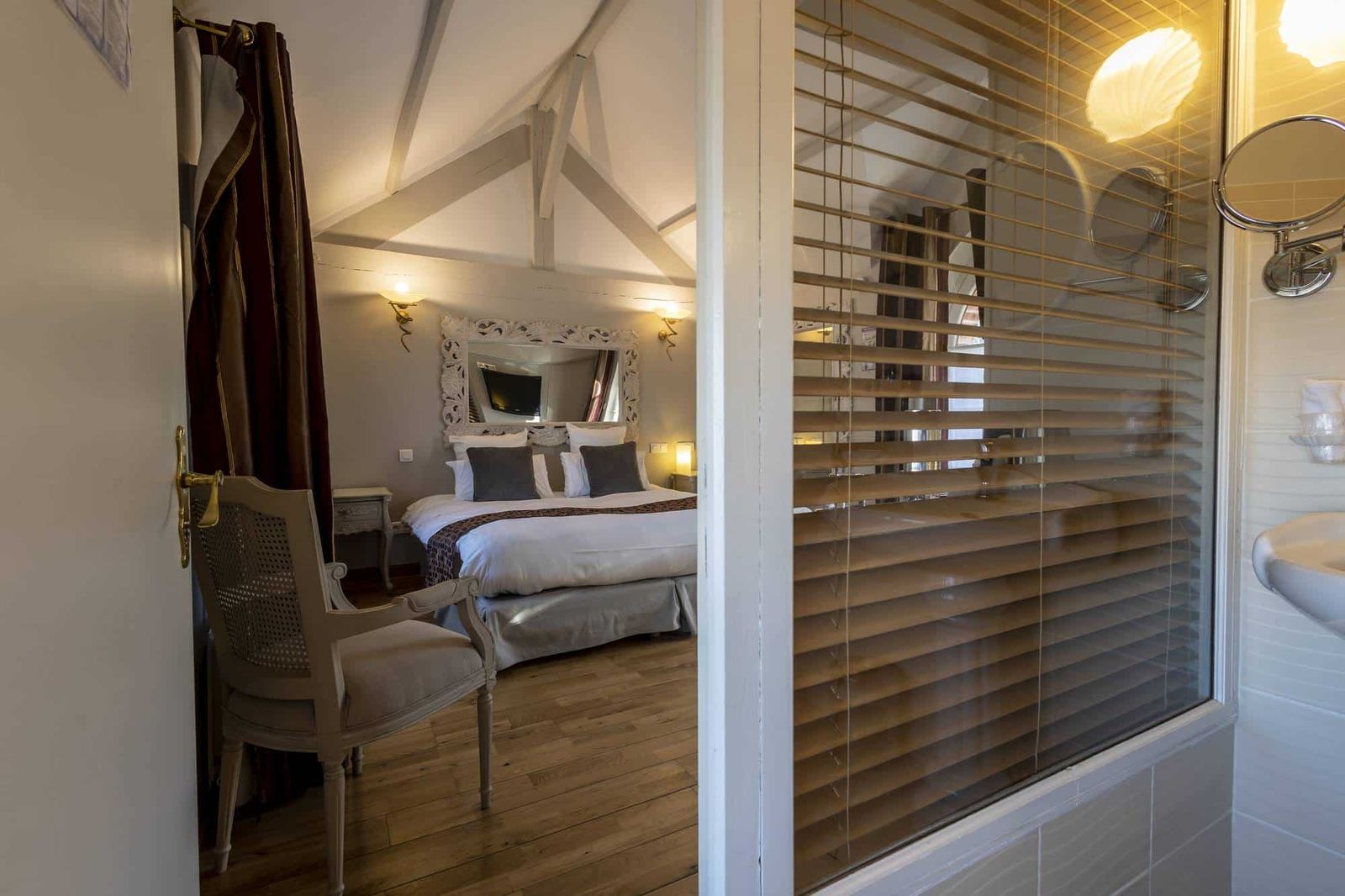 Chambre Voltige : lit sous la charpente, miroir baroque blanc, fauteuil canné et parquet