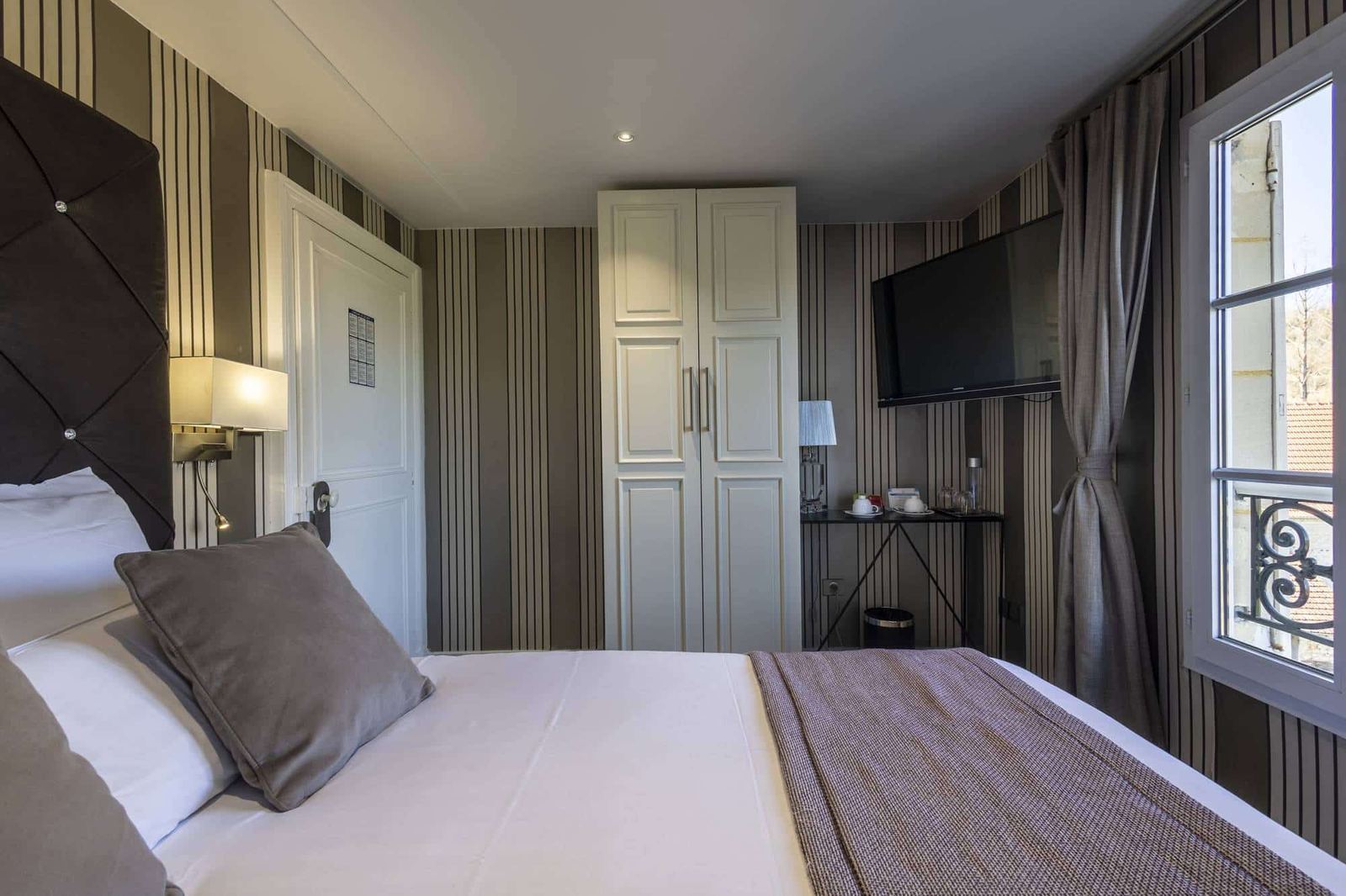 Chambre Vue sur Ferme : lit capitonné, armoire blanche et fenêtre ouverte au garde-corps en fer forgé