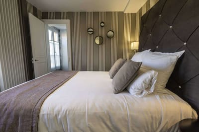 Chambre Vue sur Ferme : lit capitonné brun, coussins taupe et miroirs ronds sur papier peint rayé