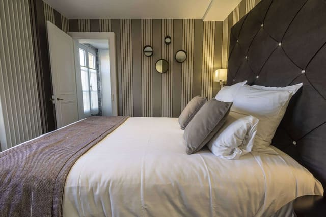 Chambre Vue sur Ferme : lit capitonné brun, coussins taupe et miroirs ronds sur papier peint rayé