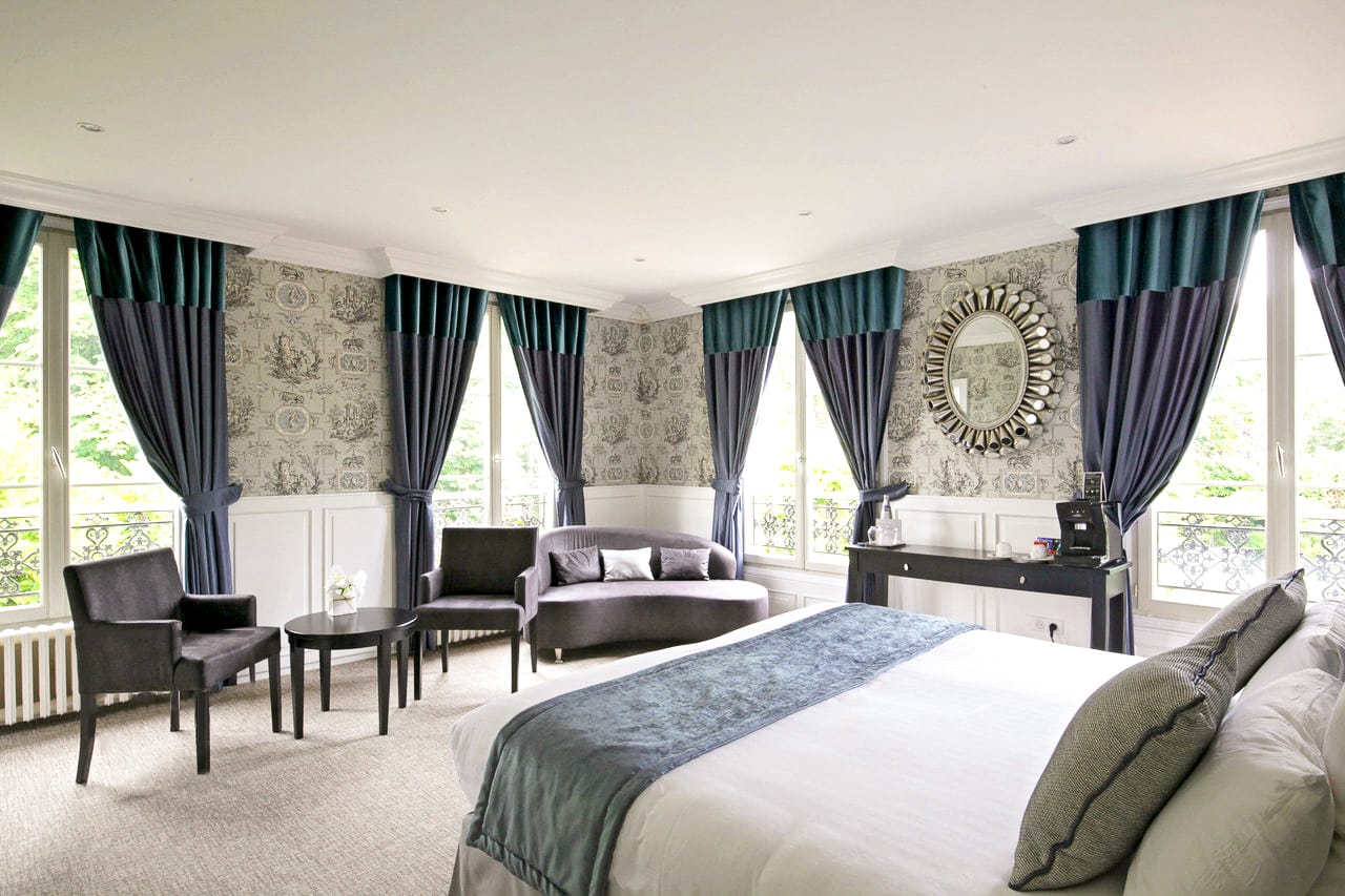 Suite Marquise : grand lit, salon aux fauteuils gris et fenêtres à balconnet sur les arbres