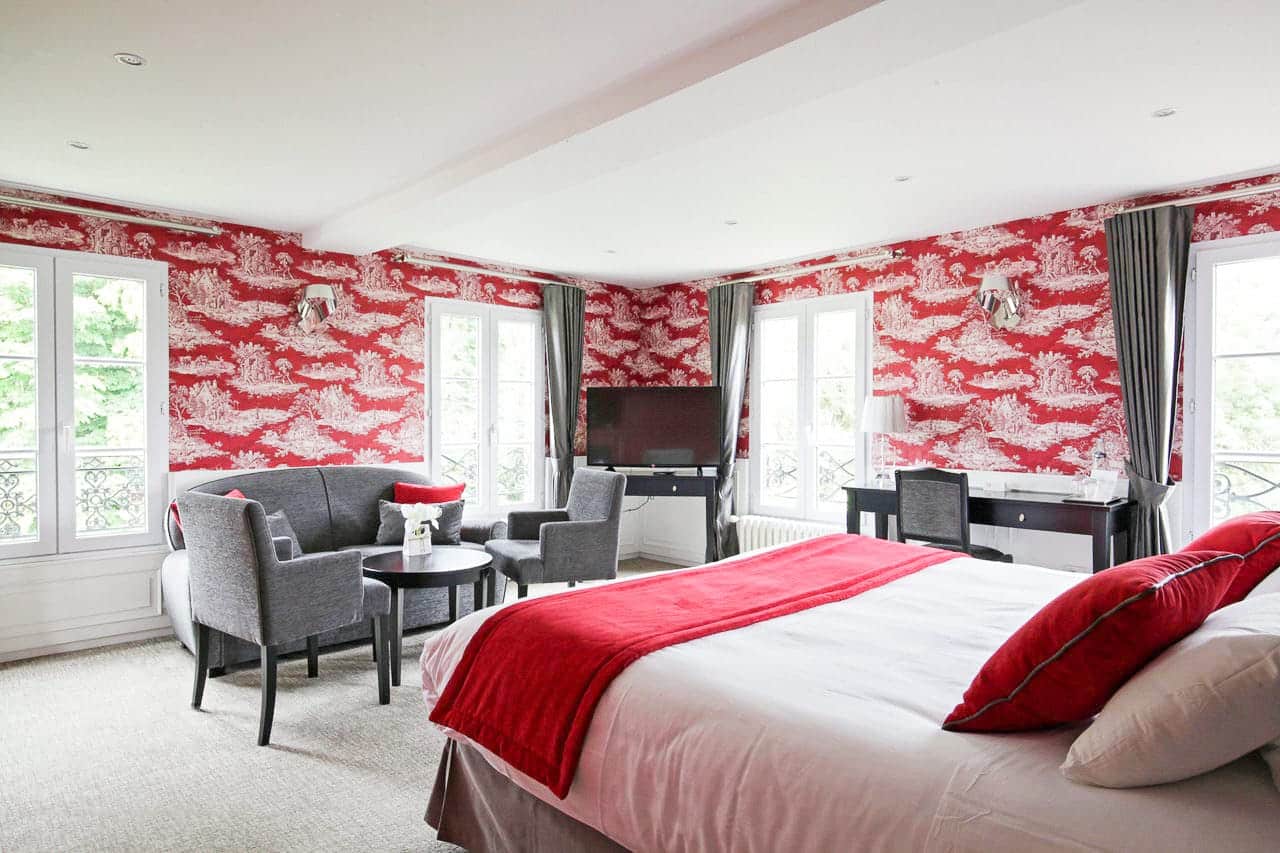 Suite Arlequin : grand lit au plaid rouge, salon gris et trois fenêtres sur la verdure