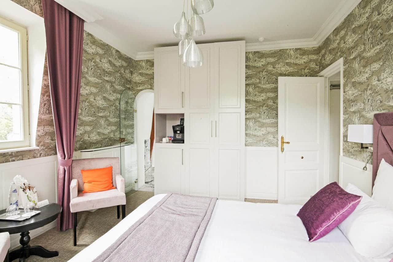 Suite Royale : armoire blanche avec machine à café, fauteuil rose près de la fenêtre et lit