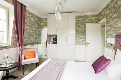 Suite Royale : armoire blanche avec machine à café, fauteuil rose près de la fenêtre et lit